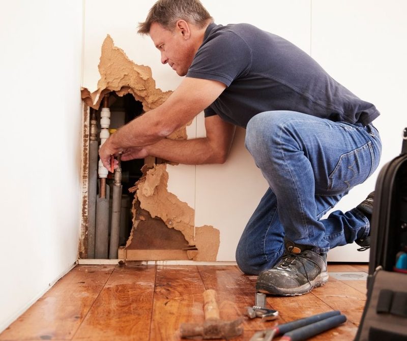 Contractor kneeling at an opened wall with exposed plumbing and broken drywall
