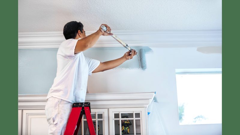 Painter on a ladder painting the top of a wall in a white room