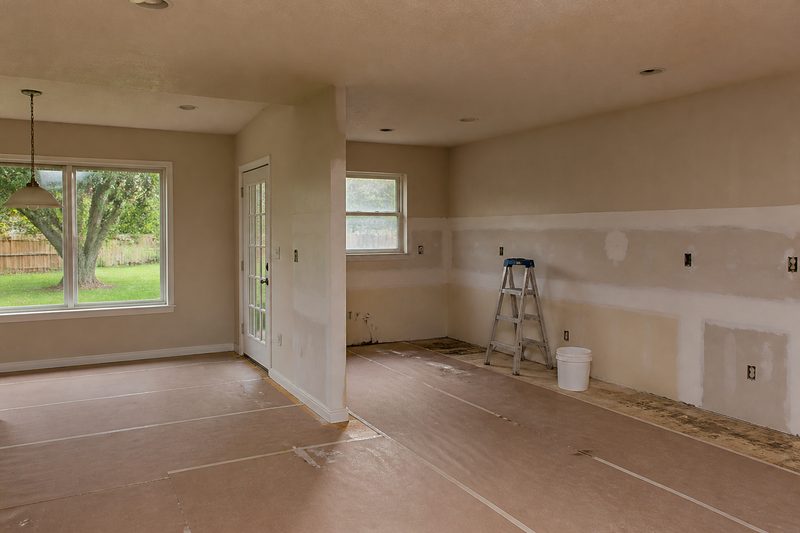 Kitchen and dining area mid-rebuild after a plumbing leak, with new drywall taped along the lower walls, cabinets removed, paper floor protection and a stepladder
