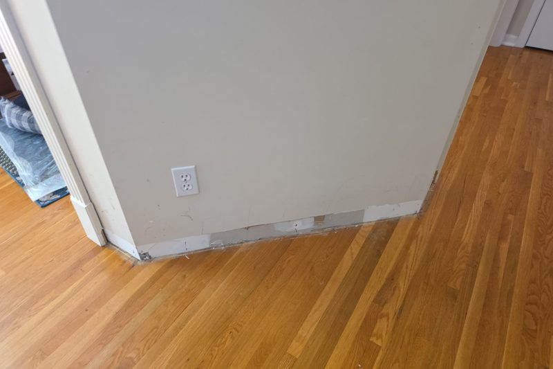 Wall corner with the baseboard removed and patched drywall along the bottom, next to oak hardwood flooring, before trim and paint